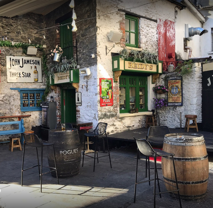 Rustic old pub exterior with barrels, chairs, green doors, and vintage wall signs