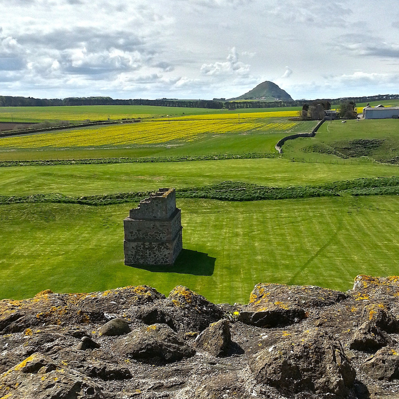 View from castle ruins over green fields with a small stone tower and cloudy sky