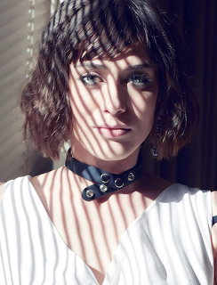 Portrait of a woman with short wavy hair in striped light, wearing a white top and black choker