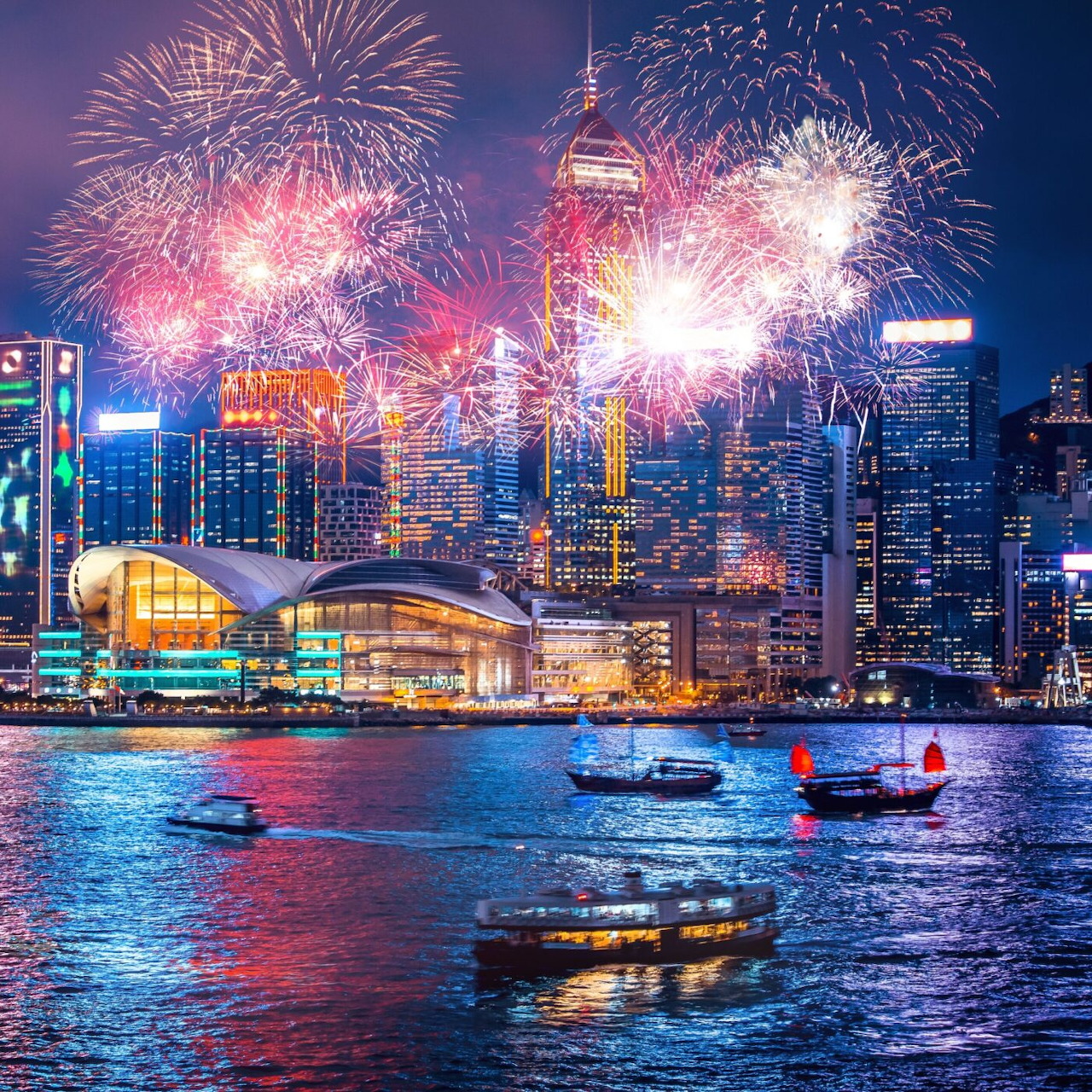 Vibrant fireworks exploding above a harbor cityscape at night with boats on water and illuminated buildings