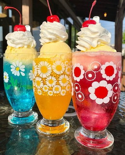 Three colorful milkshakes with whipped cream, cherries, and floral-patterned glasses in blue, orange, and red