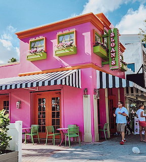 Bright pink ice cream shop building with black and white striped awnings and colorful chairs on sunny sidewalk