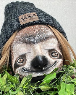 Close-up of a smiling sloth wearing a gray knit beanie hat with blonde hair and green leaves