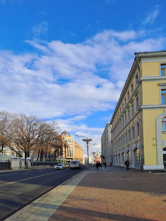 Wide city square with yellow neoclassical buildings, bare trees, paved plaza, and blue sky with white clouds