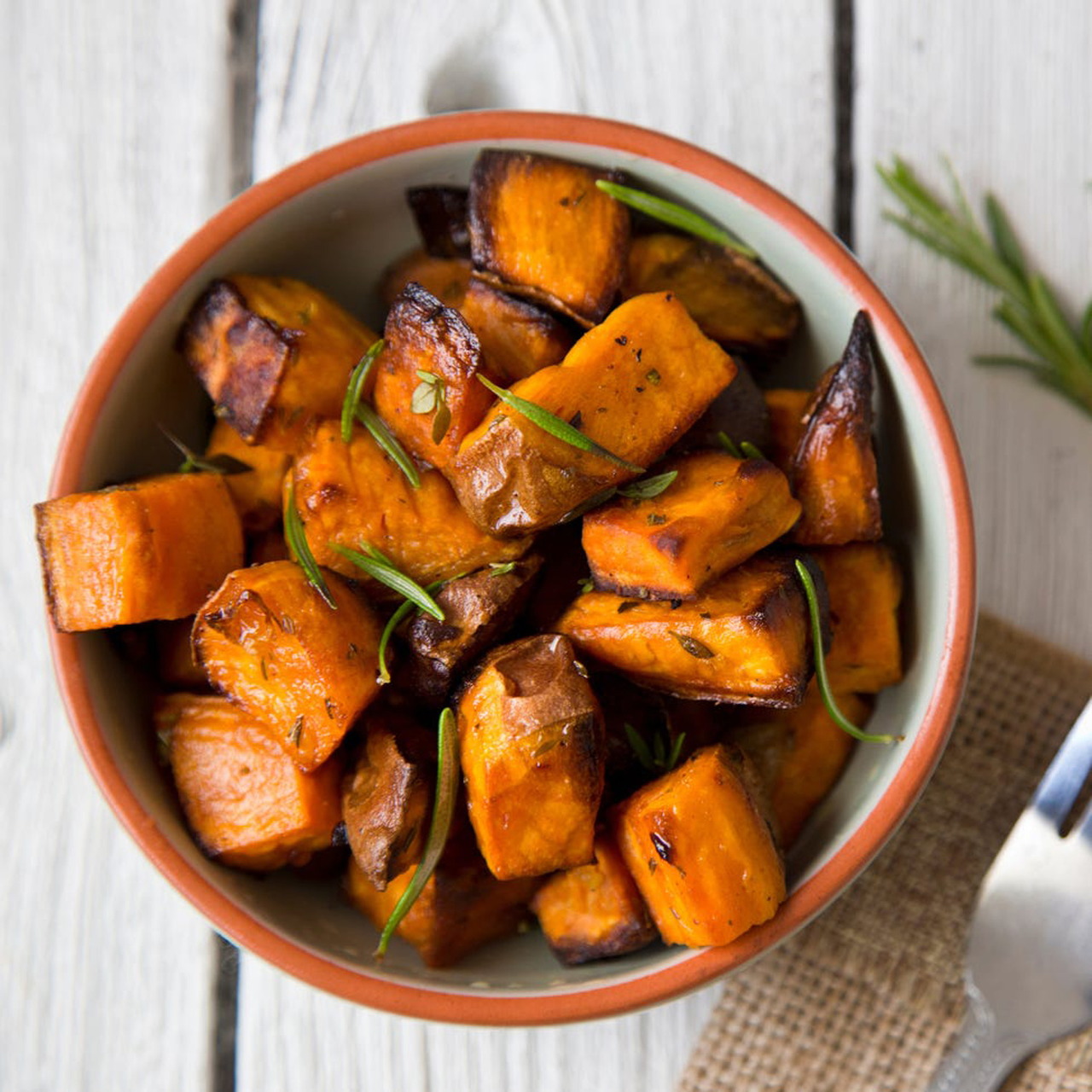 Bowl of golden roasted sweet potato chunks with herbs on white wood table