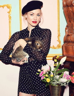 Woman in a polka dot dress and beret holding two rabbits beside flowers indoors