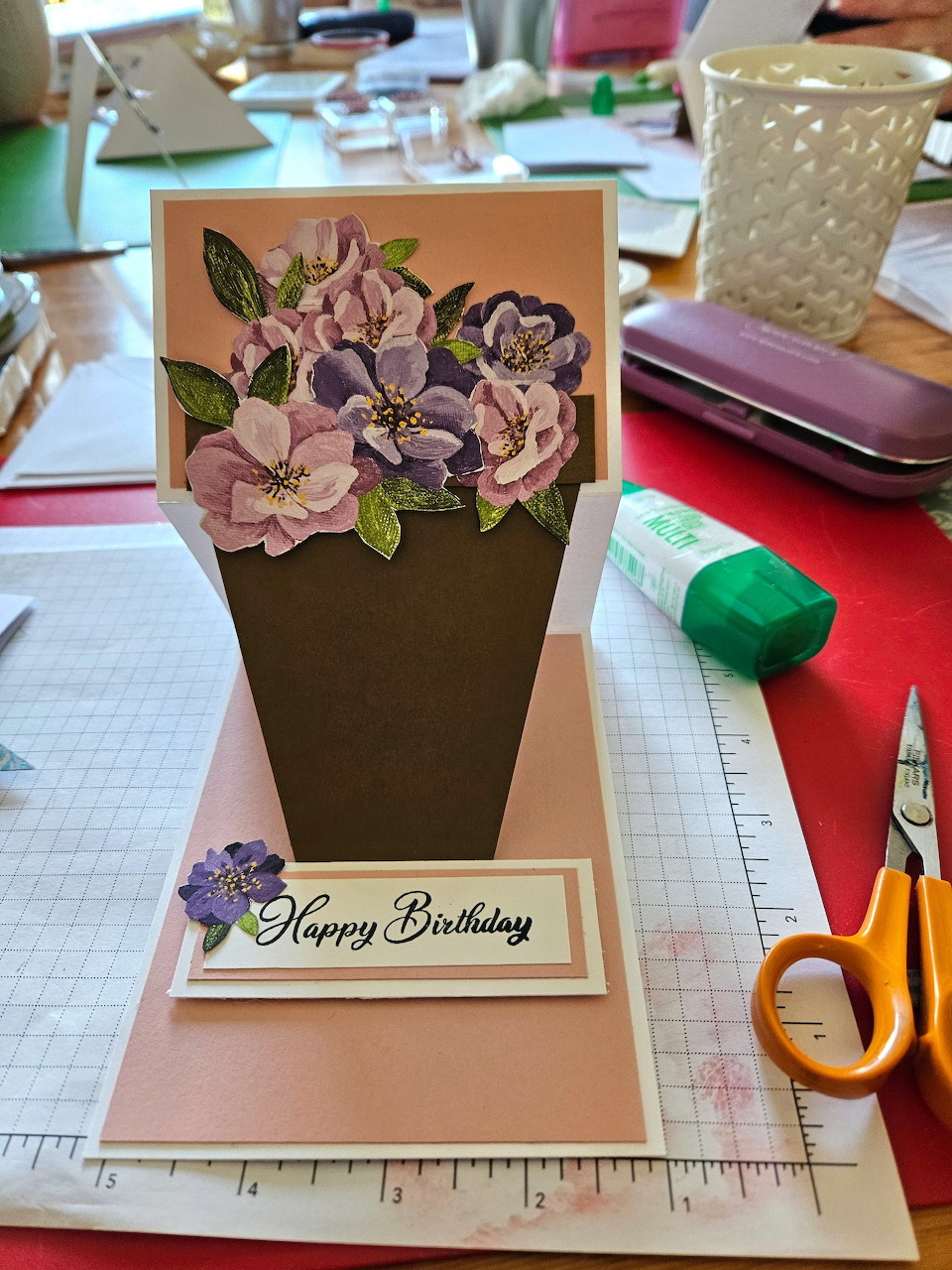 Handmade birthday card with purple flowers standing on a craft table with colorful paper and scissors