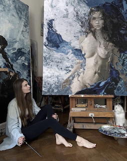 Woman artist sitting on studio floor next to large abstract figurative oil painting with white cat nearby