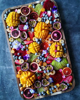Overhead view of a wooden platter with sliced mango, kiwi, berries, citrus, flowers, and colorful sweets.