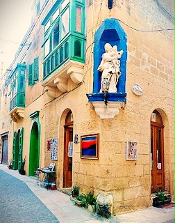 Narrow Mediterranean street with limestone buildings, green balconies, and a religious statue in a blue wall niche