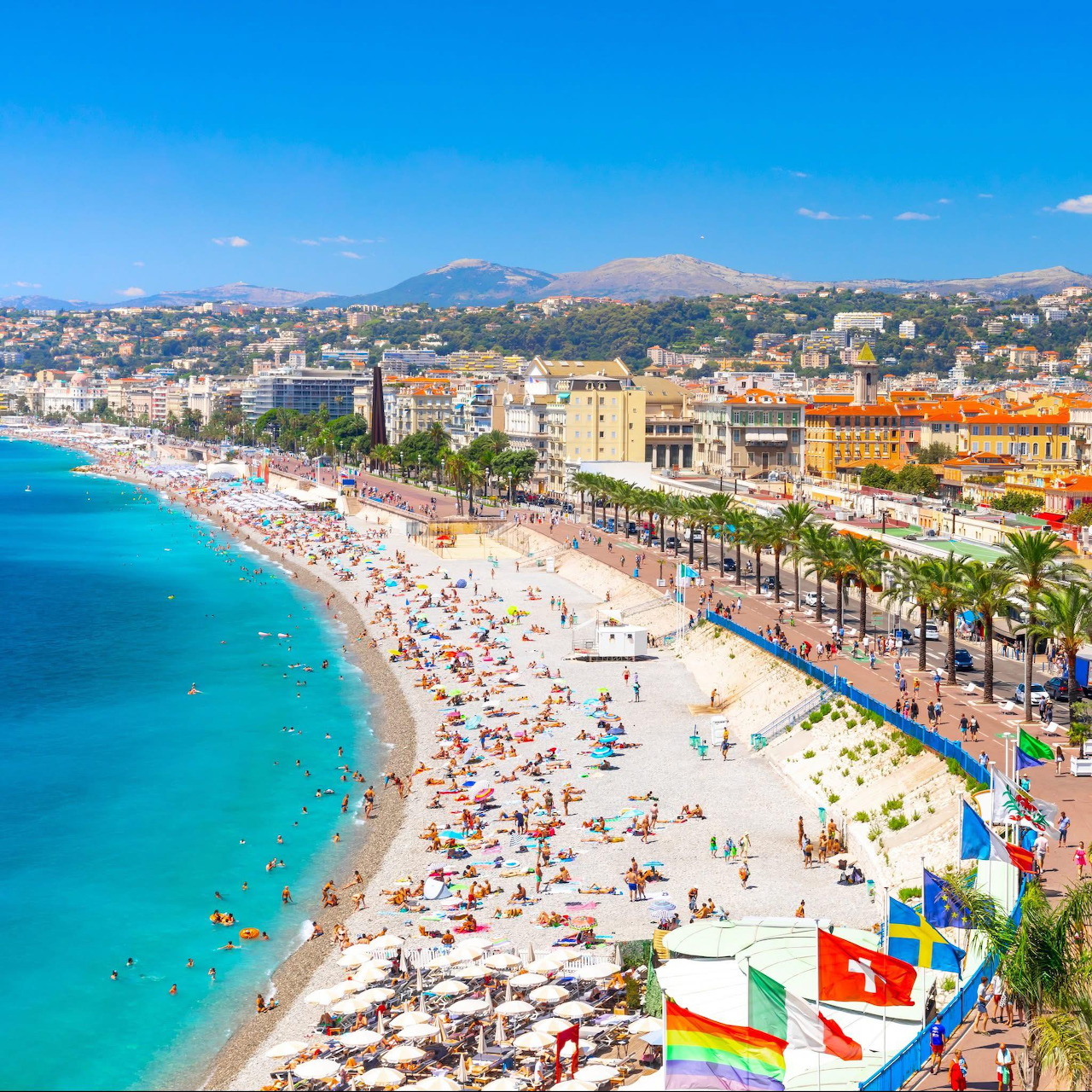 Crowded beach along Nice, France coastline with turquoise water and palm-lined promenade