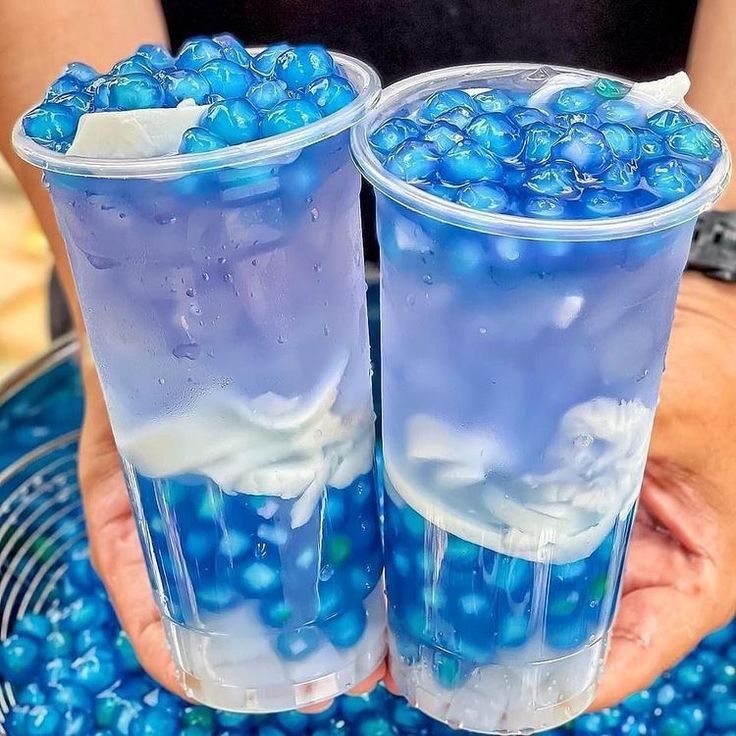 Two clear cups of blue boba tea with white cream layers held in hands