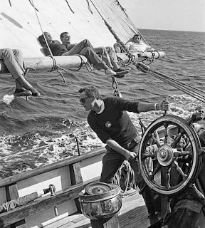 Black and white photo of sailors on a sailing ship, one steering at the wheel while others rest on the boom