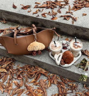 Iced chai lattes on a tray next to a tan leather bag and croissant toy surrounded by autumn leaves