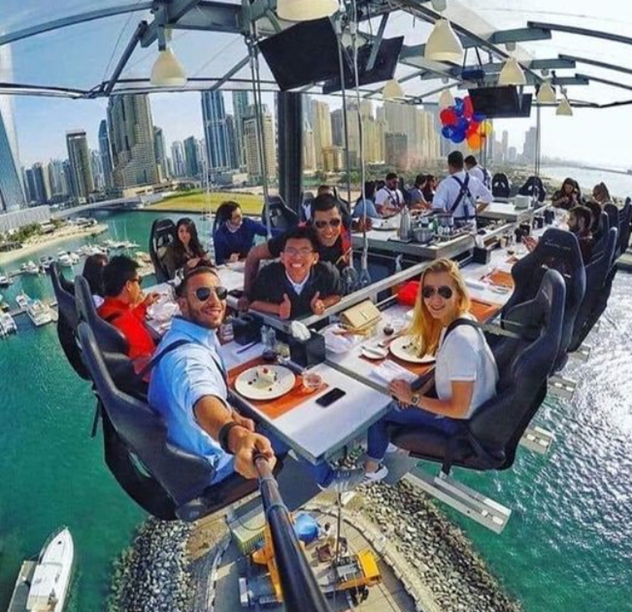 People dining at a table suspended in the air above water with city skyline in background