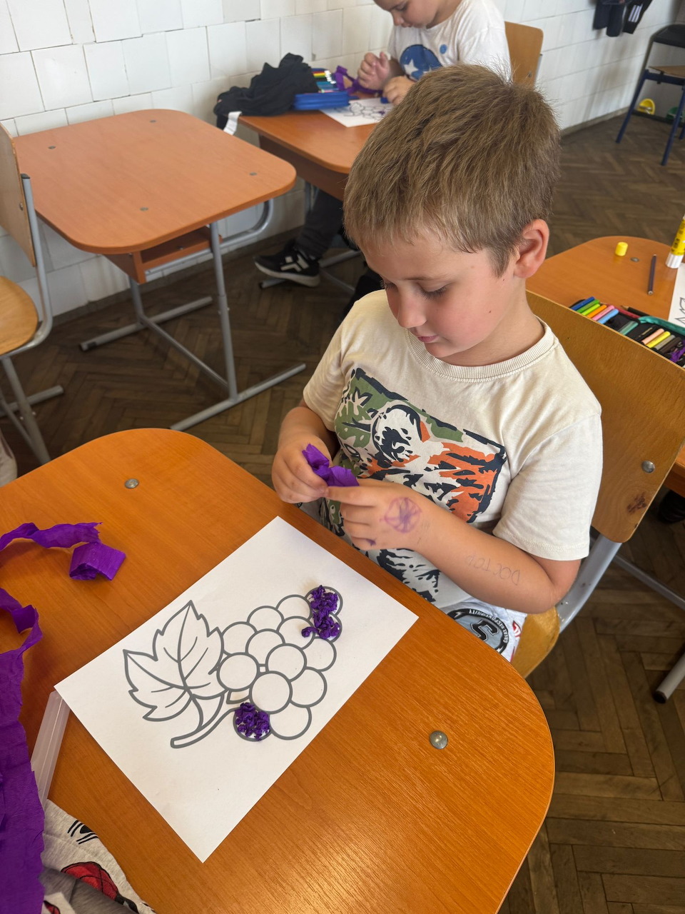 Young boy at a school desk coloring a grape cluster drawing with purple marker