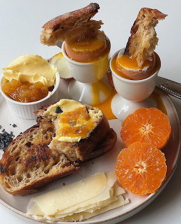 Breakfast plate with soft-boiled eggs, bread slices, cheese, jam, and fresh orange slices on white dishes