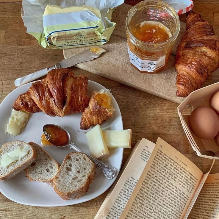 Croissants, bread, butter, jam, eggs, and an open book on a wooden table