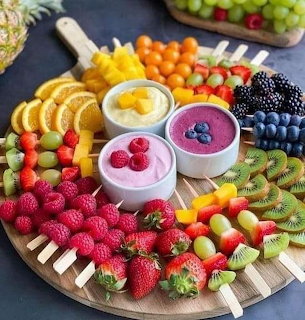 Round wooden board with colorful fruit skewers and three bowls of yogurt dipping sauces