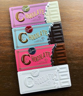 Four decorative chocolate bars in purple, blue, pink and white arranged vertically on wooden table