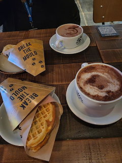 Two cups of coffee with latte art and waffles in paper wrapping on a wooden café table table