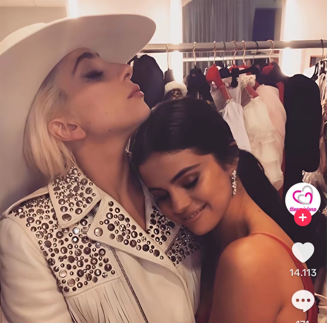 Two women posing closely backstage beside a clothing rack