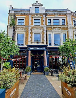 Ornate yellow brick Victorian pub building with white trim, outdoor seating, and potted plants in front