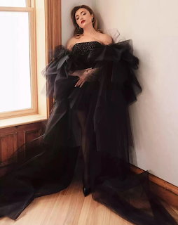 Woman in dramatic black tulle strapless gown posing against a wall near a window