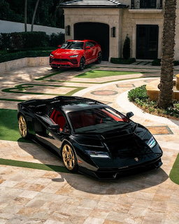 Black Lamborghini coupe and red Lamborghini SUV parked on a stone driveway outside a large house