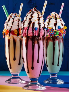 Three colorful milkshakes with whipped cream, chocolate sauce, and sprinkles in tall glass cups with striped straws