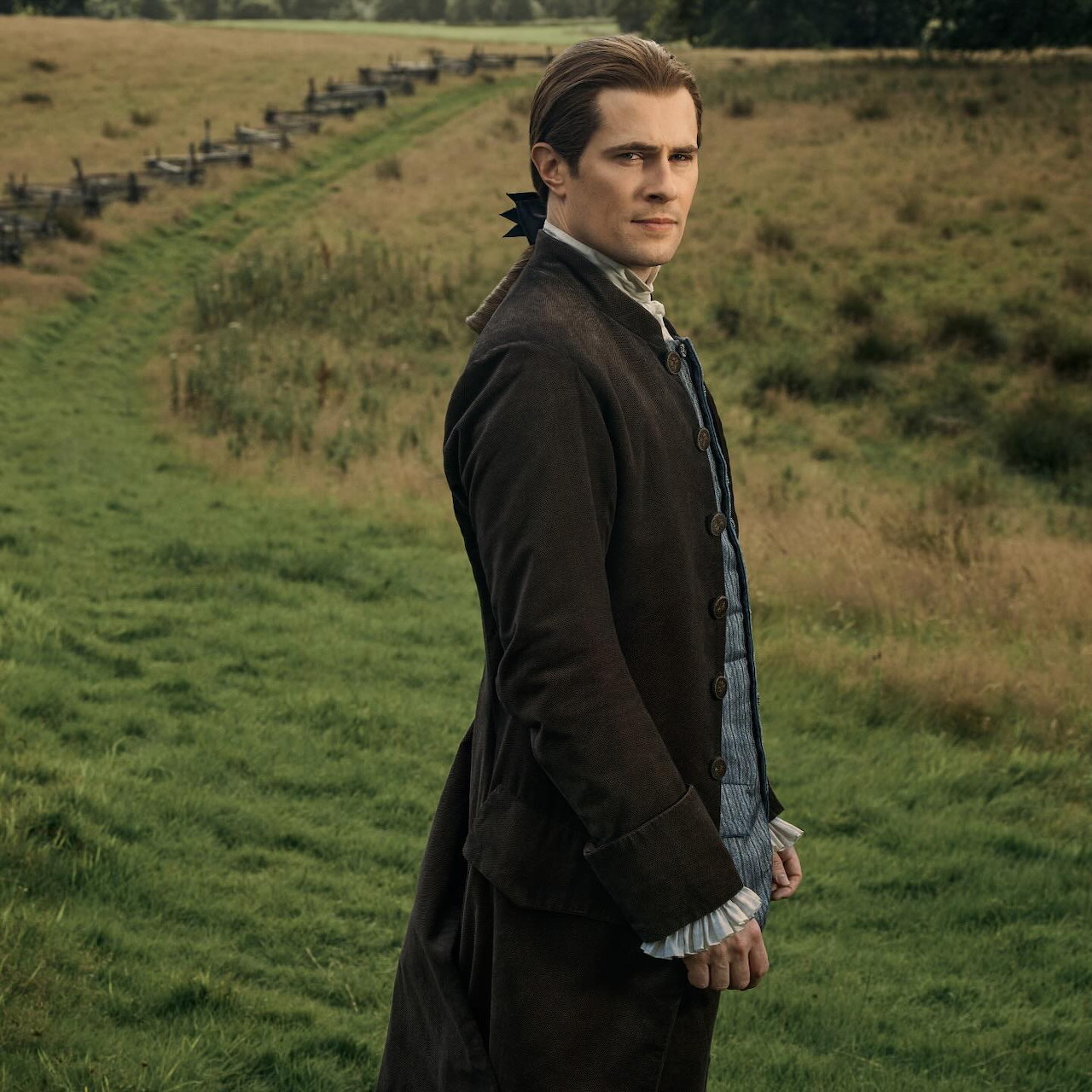 Man in dark period clothing standing on a grassy path in a field