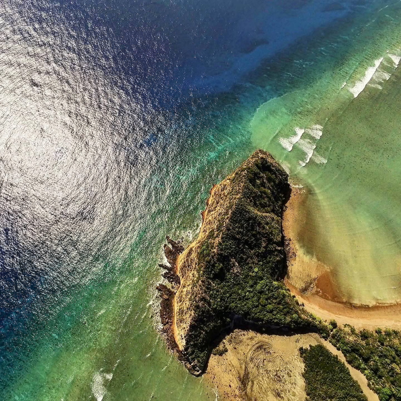 Aerial view of a rocky coastal outcrop, sandy beach, and clear blue-green ocean