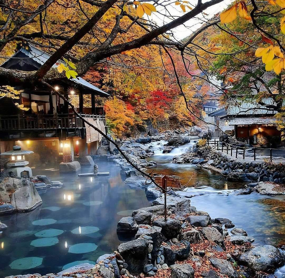 Outdoor hot spring pools beside autumn trees and mountain stream with traditional Japanese buildings