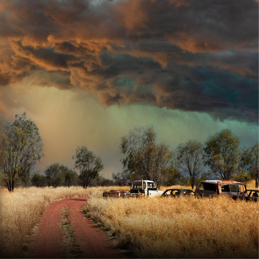 Dark storm clouds over a dirt road with dry grass, trees, and old vehicles