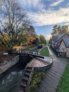Canal lock with wooden gates, autumn trees, brick cottage, and green towpath under blue sky