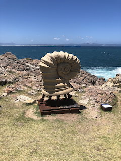 Large shell sculpture on rock base beside ocean cliffs with blue water and sky