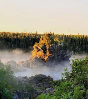 Large rocky outcrop emerging from mist over a river surrounded by green boreal forest at sunrise