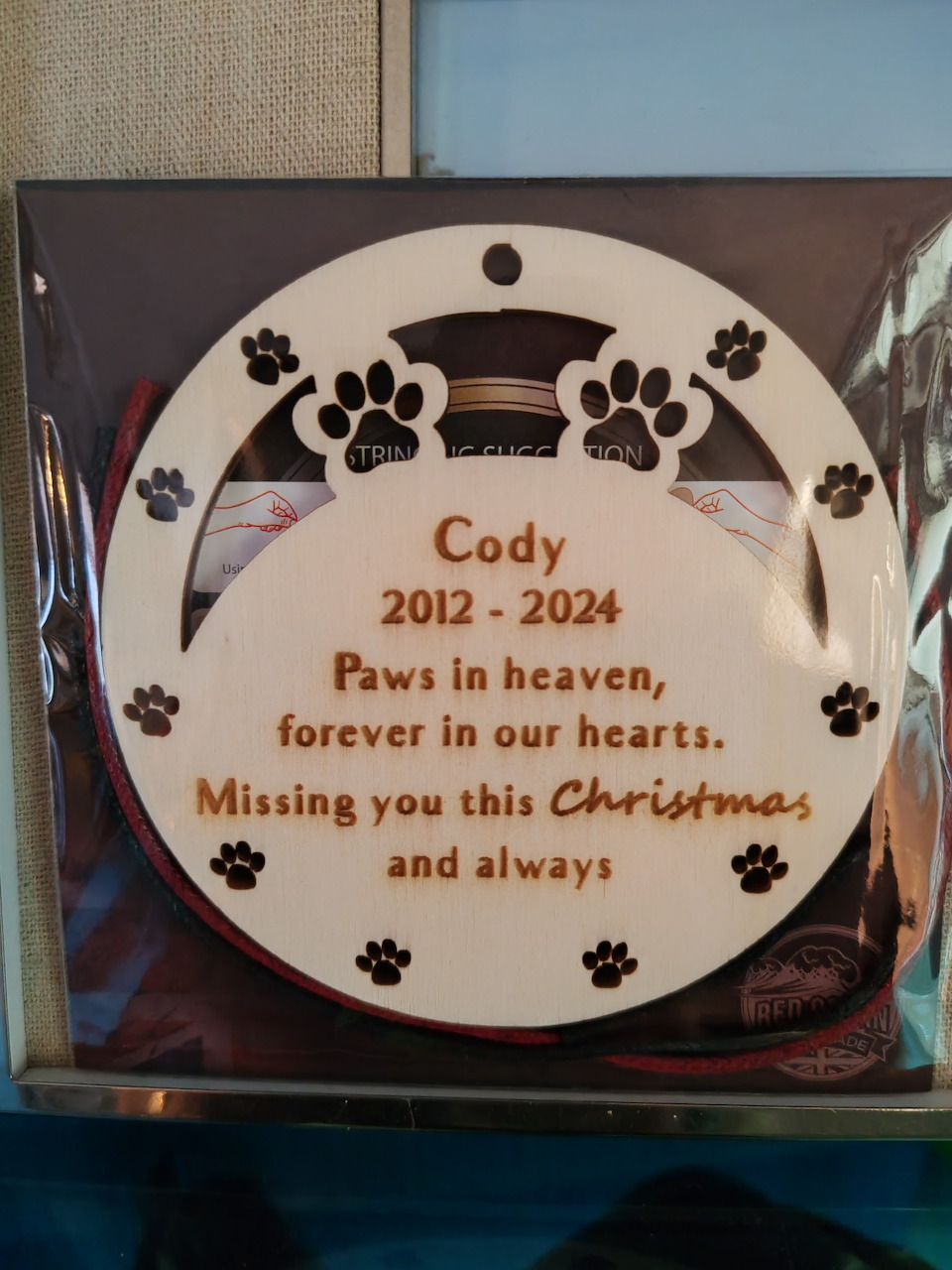White circular memorial ornament with black paw prints and red text reading Cody 2012-2024