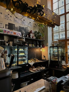 Cafe interior with coffee counter, pastry displays, shelves, plants, and tall windows