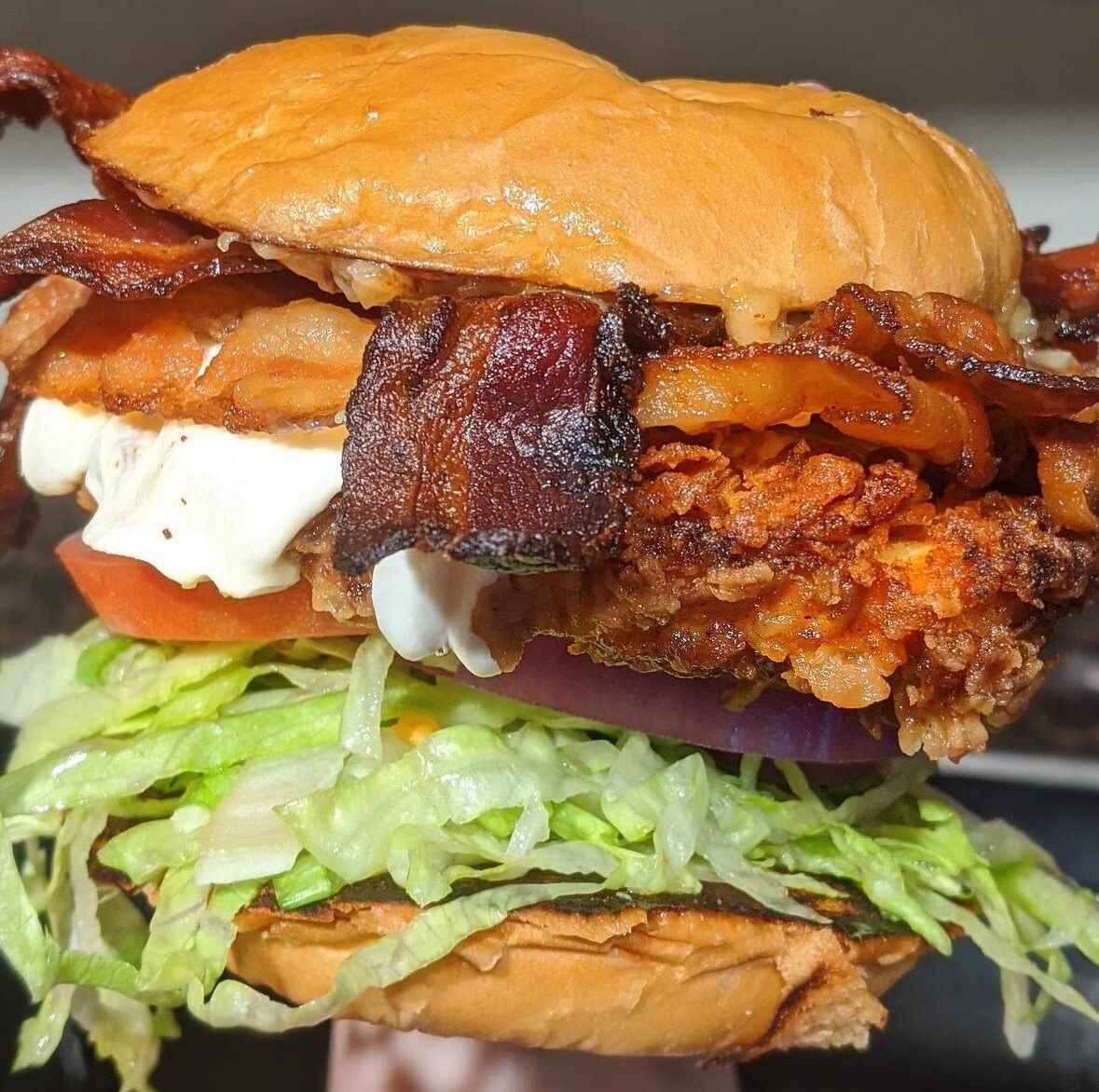Close-up of a fried chicken sandwich with bacon, lettuce, tomato, onion, and sauce