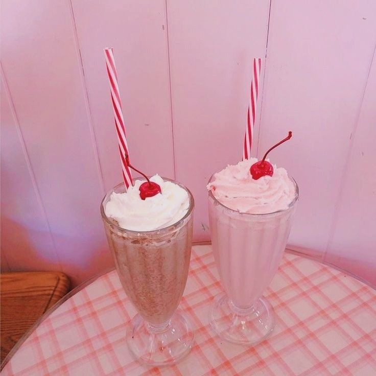 Two milkshakes topped with whipped cream and cherries on a pink table