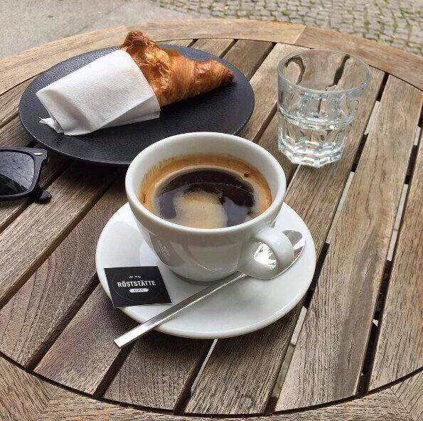 Black coffee and a croissant on a wooden café table with a water glass and sunglasses