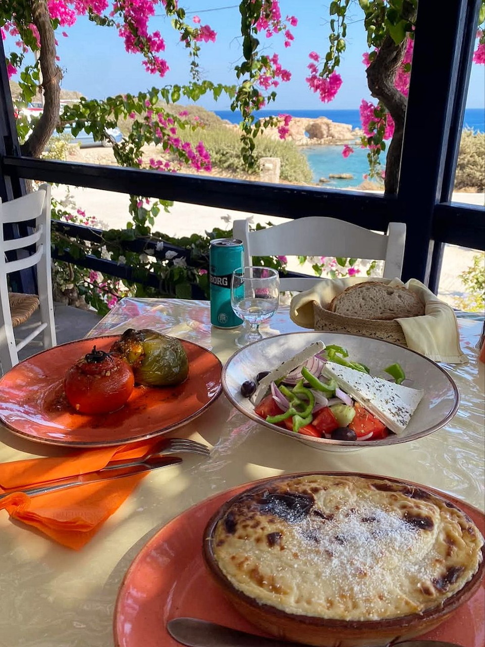 Outdoor terrace table with Mediterranean dishes including stuffed peppers, salad, and baked casserole, with sea view and pink flowers.