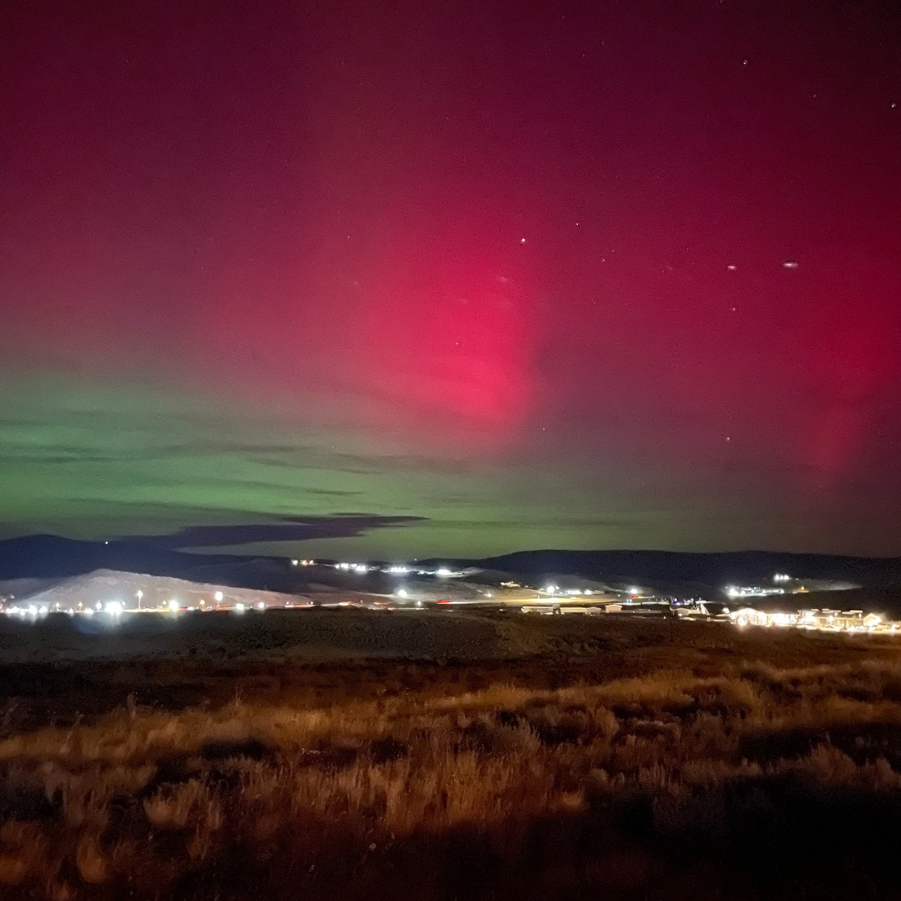 Red and green aurora over a dark grassy landscape with distant lights at night
