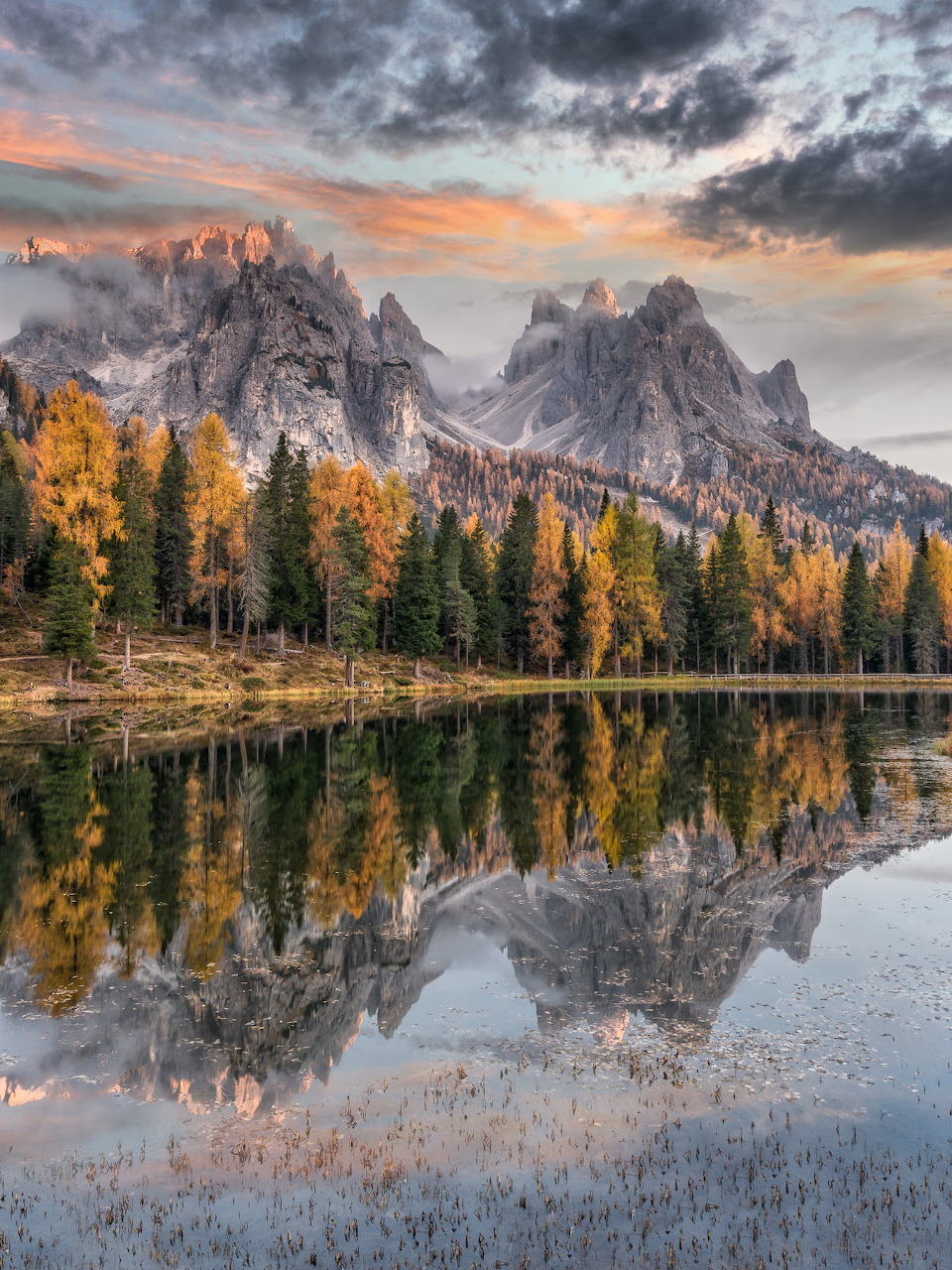 Lago Antorno reflecting Dolomite peaks and autumn trees under dramatic clouds at sunset