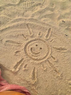 Smiling sun with rays drawn in beach sand, pink fabric visible at bottom left