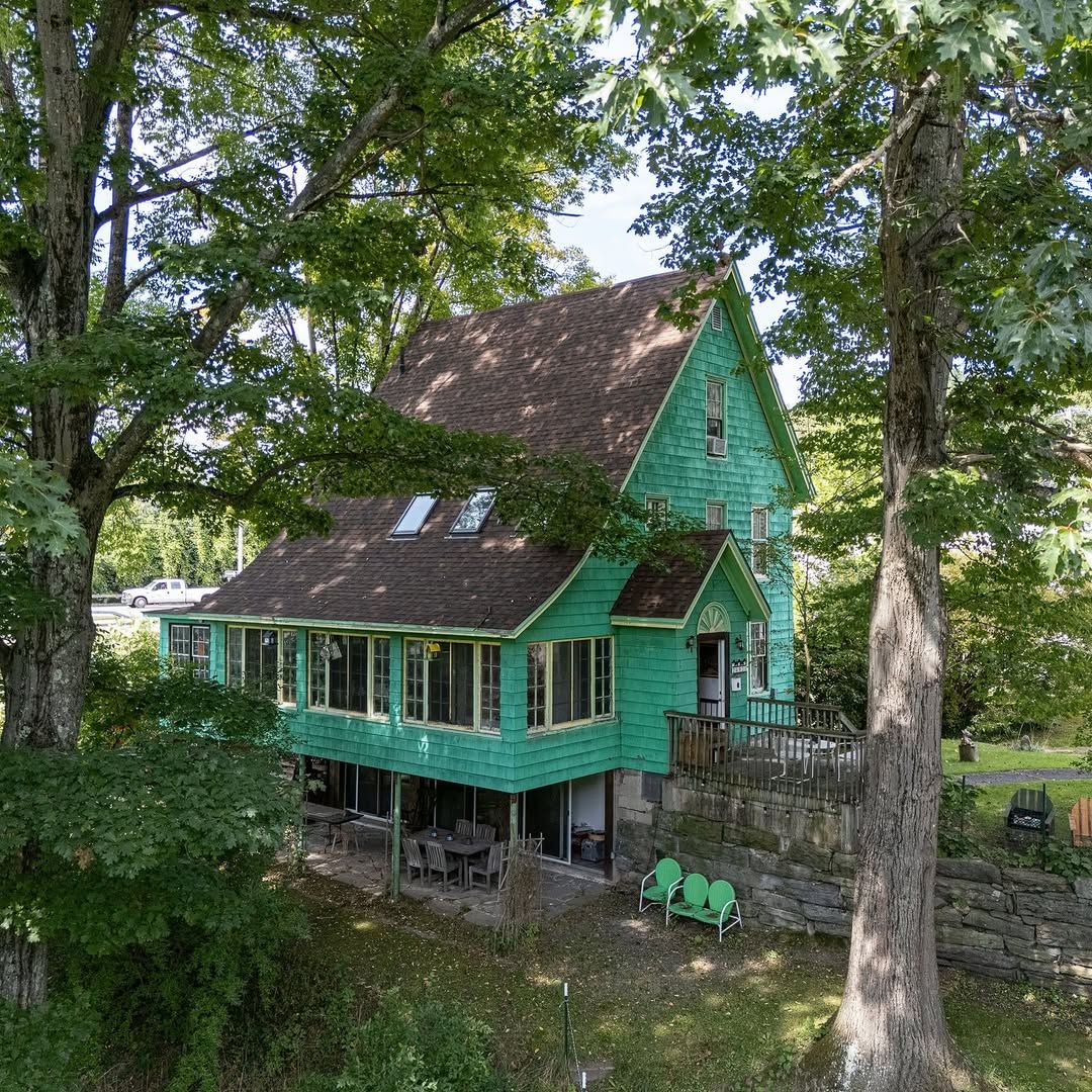 Green two-story wooden house with deck surrounded by large trees