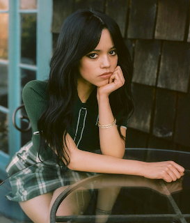 Jenna Ortega sitting at a glass table, resting her face on one hand