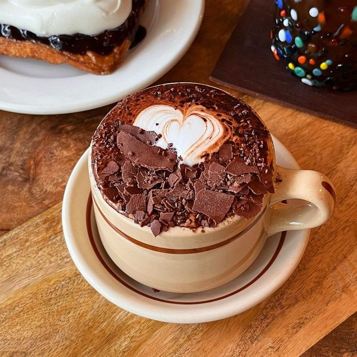 Chocolate mocha with whipped cream and chocolate shavings in beige cup on wooden surface with s'more beside it