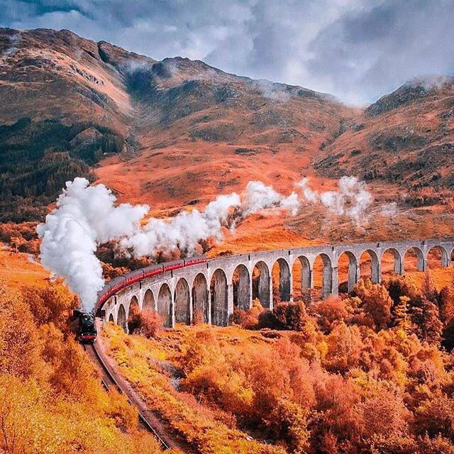 Steam train crossing stone arched viaduct through orange autumn landscape with mountains